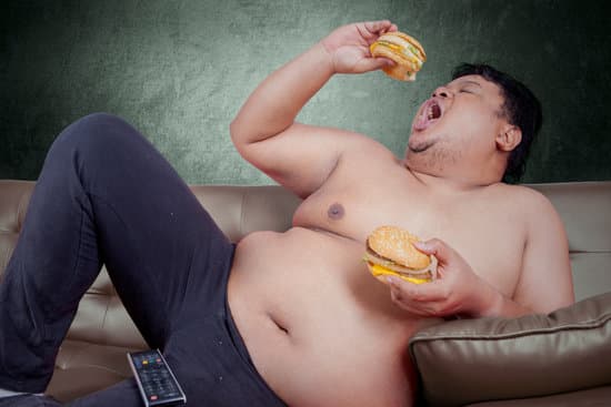 Picture of obese person looks hungry and eat two hamburger while leaning on the sofa